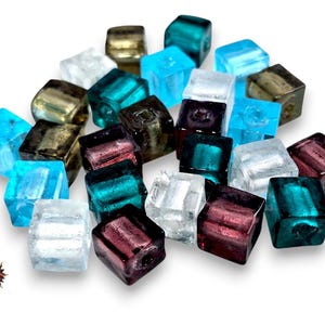 Può includere: Una collezione di piccole perline di vetro di forma quadrata in vari colori traslucidi, tra cui verde acqua, trasparente, viola scuro e bronzo metallizzato. Ogni perlina presenta un foro centrale per l'infilatura.
