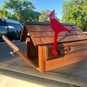 May include: A wooden birdhouse featuring a red cardinal silhouette. The birdhouse has a brown shingled roof and a hinged door with a silver knob. The wood grain is visible on the front and sides of the birdhouse.