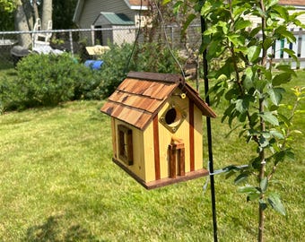May include: A wooden birdhouse with a light yellow body and dark brown vertical trim, featuring a shingled roof, a circular entrance hole with a brass plate, a small wooden perch, and a side window, suspended from a black metal shepherd's hook.