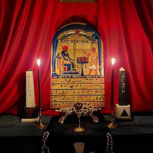 Może przedstawiać: Ciemnoczerwony aksamitny zasłonę w tle ze złotym obrazem w ramie nad drewnianym ołtarzem. Ołtarz ma dwie czarne świece, dwie białe świece i złoty metalowy stojak z okrągłym blatem i trzema nogami. Ołtarz ma również drewnianą tabliczkę z hieroglifami i przedstawieniem dwóch postaci. Tabliczka jest oznaczona jako "OGHAM" i "ALATH".