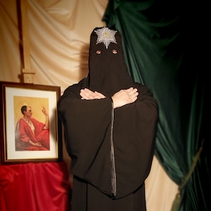 May include: A person wearing a black hooded robe with a white star design on the hood. The person is standing in front of a red and green fabric background.