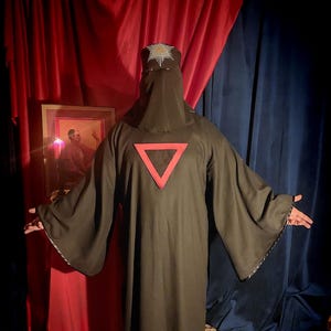 May include: A dark robe with a red triangle on the back. The robe has wide sleeves and a hood that covers the head and face. The hood is adorned with a decorative emblem. The background includes red and blue curtains and a framed picture.
