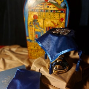 May include: A blue velvet cushion with a gold base and a decorative silver design on top. The cushion is in front of a tall, rectangular, painted wooden object with hieroglyphics.