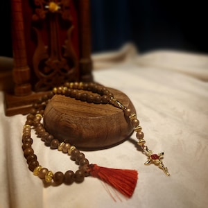 May include: A wooden rosary with brown beads and gold accents, including a gold cross with red accents. A red tassel hangs from the rosary. In the background is a wooden carved cross.