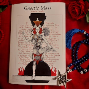 Könnte beinhalten: Ein weißes Buch mit dem Titel Gnostic Mass von Aleister Crowley, das eine zentrale Illustration einer mehrköpfigen Figur mit Löwe und Schlange vor roten Flammen und Text zeigt. Eine blaue Perlenkette mit einem silbernen Pentagramm-Anhänger liegt daneben.