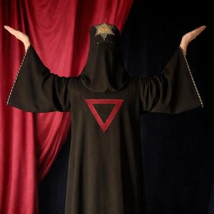 May include: Black hooded robe with a red triangle and purple square accents. The robe has wide sleeves with white and gold trim. A gold starburst design adorns the hood. The robe is worn over a patterned rug.