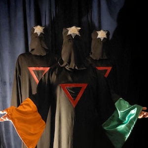 May include: Three figures in black hooded robes with red triangle emblems. The robes have orange and green sleeves and a purple patch. Each hood is adorned with a star-shaped decoration. The figures are standing in front of a dark curtain.