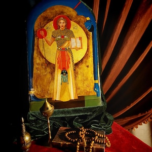 May include: A gold-leaf icon depicting a female figure in a yellow robe, holding a book and a staff. The icon is framed in a blue and green arch and set against a red velvet background. There is a small incense burner and a book of prayers with a rosary on top.