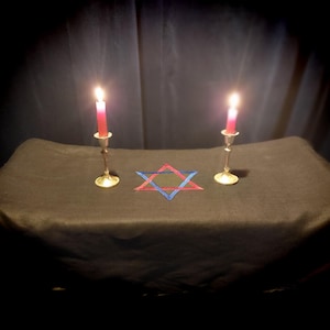 Puede incluir: Dos velas encendidas en candelabros de plata con una estrella de David azul y roja sobre un mantel negro.