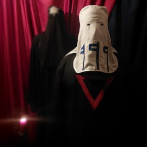 May include: A white hood with the number "199" in blue, and a red triangle on a black garment. A figure in a black garment is in the background, with a red curtain backdrop.