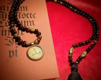 May include: A long necklace featuring alternating black and brown beads, accented with ornate gold-toned metal spacers. The necklace includes a circular gold-toned pendant with an engraved symbol and terminates in a black fabric tassel.