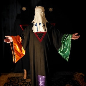 May include: A person in a black robe with orange and green sleeves, a white hood, and a red triangle. The hood has blue and black markings. The background is dark with two star-shaped lights.