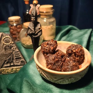 Puede incluir: Un cuenco pequeño, redondo y de color claro con patrones tallados contiene varios alimentos de color marrón oscuro, texturizados y de forma irregular. Al fondo, hay una estatua negra de una figura egipcia, una placa de piedra y frascos de vidrio.