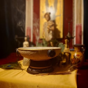 Può includere: Un bruciatore di incenso beige con una base scura, che emette volute di fumo. Il bruciatore ha un bordo strutturato e dettagli metallici. La base ha testo greco. Una piuma di pavone e altri oggetti decorativi sono nelle vicinanze.
