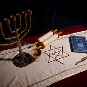 Pode incluir: Um candelabro de latão de sete braços, um pequeno pergaminho, um livro azul e um ponteiro de metal repousam sobre um caminho de mesa branco. O tecido apresenta um hexagrama vermelho bordado com uma estrela central numa superfície escura.