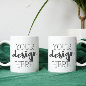 May include: Two white ceramic mugs with black text that reads "Your design here" on a green towel.