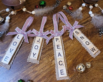 May include: Four Christmas decorations made from burlap and Scrabble tiles. The tiles spell out the words "LOVE", "JOY", "PEACE", and "SNOW". Each decoration is tied with a red and white gingham ribbon.
