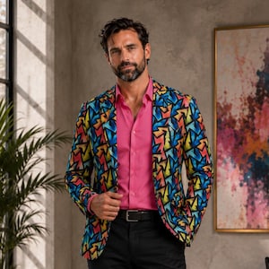 Op de afbeelding: Een blazer met een levendig, meerkleurig geometrisch patroon in blauwe, gele en rode tinten, gedragen over een effen roze overhemd met kraag, gecombineerd met een zwarte broek en een zwarte riem met een zilveren gesp.