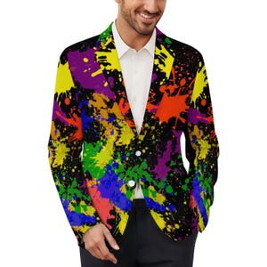 Puede incluir: Un blazer vibrante con un diseño de salpicaduras de pintura coloridas en amarillo, morado, naranja, azul y verde sobre un fondo negro. El blazer tiene un cierre clásico de dos botones y una camisa blanca debajo.