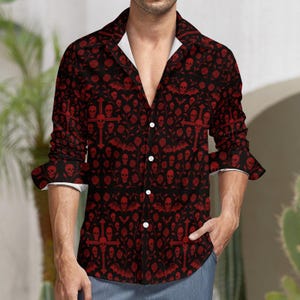 Peut inclure: Une chemise boutonnée noire avec un motif répété de crânes rouges, de croix, de chauves-souris et de roses. La chemise a un col et des poignets blancs. Les manches sont retroussées et le haut est déboutonné.