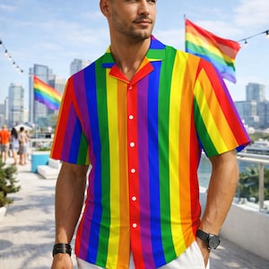 May include: A short-sleeved button-down shirt with vertical rainbow stripes. The shirt has a camp collar and white buttons. The person is wearing white shorts and a black watch. Rainbow flags are visible in the background.
