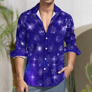 Op de afbeelding: Een lang mouw overhemd met knopen en een galaxy print met sterren, manen en andere hemellichamen in wit op een donkerblauwe achtergrond.