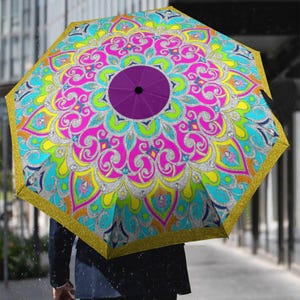 May include: A colourful umbrella with a mandala design. The umbrella has a pattern of pink, blue, yellow, and green, with a purple centre and gold trim. The umbrella is open, providing shelter from the rain.