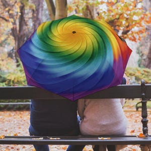 Könnte beinhalten: Ein farbenfroher Regenschirm mit einem Regenbogen-Spiralmuster. Das Muster des Regenschirms weist einen Farbverlauf auf, darunter Blau, Lila, Grün, Gelb und Rot. Der Regenschirm ist geöffnet, und das Bild zeigt die Oberseite des Regenschirms.