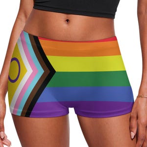 May include: A pair of colorful shorts featuring the Progress Pride flag design. The shorts have a rainbow stripe pattern with additional colors and symbols representing various LGBTQ+ identities. The shorts are a vibrant mix of colors.