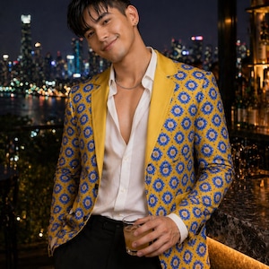May include: A yellow blazer with a blue floral pattern, paired with a white shirt and black pants. The blazer has a yellow collar and lapels. The person is holding a glass of a brown beverage. The background features a city skyline at night.