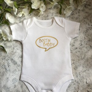 May include: White baby grow with a gold speech bubble that says "BOSTIN BABBY".