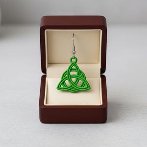 May include: A single green Celtic knot earring displayed in a dark brown jewelry box with a cream-colored interior. The earring is a detailed, three-part design, suspended from a silver-colored hook. The box is open, showcasing the earring against a soft background.