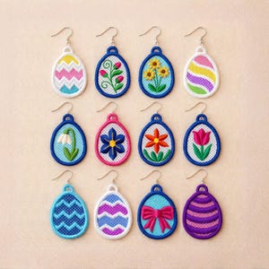 Pode incluir: Uma coleção de brincos bordados em forma de ovo de Páscoa com vários desenhos, incluindo flores, ziguezagues e laços. Cada brinco tem um gancho prateado e uma borda azul ou branca, com detalhes coloridos.