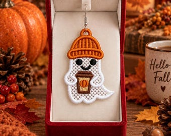 May include: A white embroidered ghost earring featuring a textured orange beanie and holding a brown coffee cup with a small orange pumpkin icon. The ghost has black circular eyes and a curved black mouth, presented in a red velvet jewelry box.