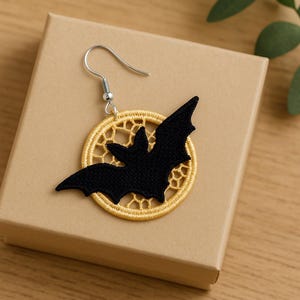 May include: A black bat-shaped earring with a gold-coloured, circular embroidered frame. The earring has a silver-coloured hook. The earring is displayed on a tan-coloured box. The earring is a Halloween-themed accessory.