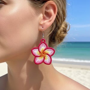 Puede incluir: Pendientes bordados con forma de flor en una vibrante combinación de colores rosa, blanco y amarillo. Los pendientes tienen forma de flor de plumeria, con un diseño de puntadas detallado. Los pendientes cuelgan de un gancho plateado. El fondo es una escena de playa con agua y cielo azules.