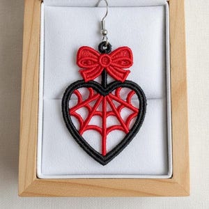 May include: An embroidered earring featuring a red heart shape with a black border and a red spiderweb pattern inside. A red bow is attached to the top of the heart, which connects to a silver-toned metal hook for hanging the earring.