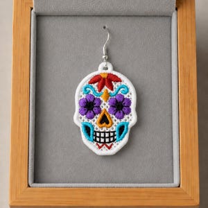 May include: A single earring featuring a white skull-shaped pendant with intricate embroidery. The design includes a red floral detail at the top, purple flower-shaped eye sockets, a yellow nose, and a black grid-patterned mouth with blue accents.