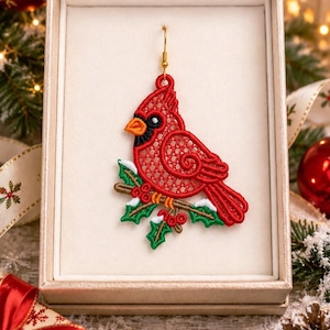 May include: A red cardinal bird earring featuring intricate lace-like embroidery, perched on a brown branch with green holly leaves and red berries. The ornament is suspended from a gold-toned metal hook and presented inside a beige gift box.