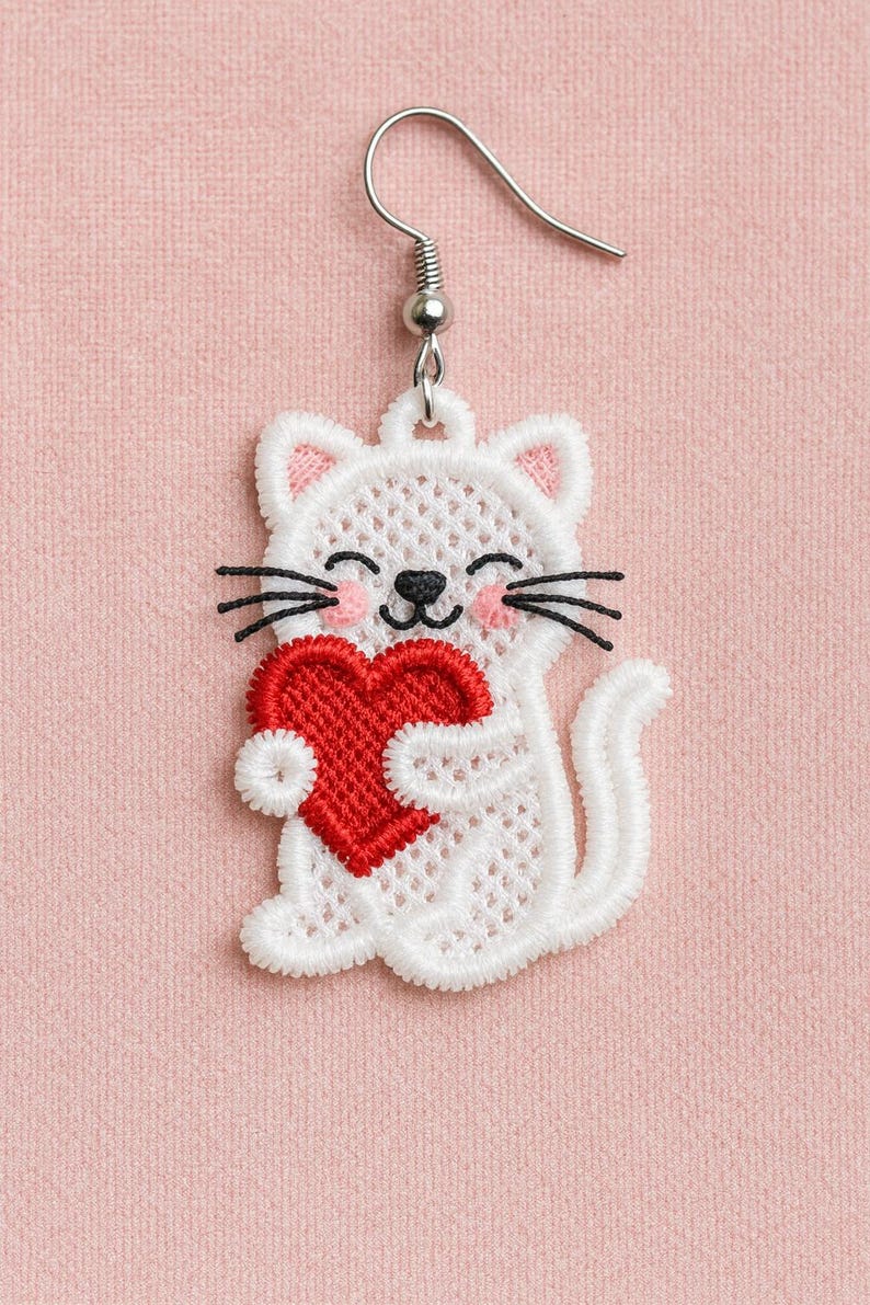 Op de afbeelding: Witte katvormige oorbel met een rood hart. De kat heeft zwarte snorharen, roze wangen en roze binnenoren. De oorbel is gemaakt van kant en heeft een zilverkleurige haak.