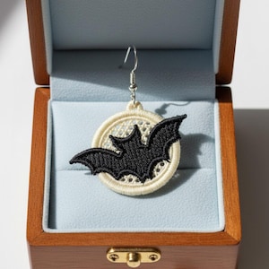May include: A single earring featuring a black embroidered bat centered on a circular cream-colored mesh background. The earring is attached to a silver-toned metal hook and is displayed inside a wooden box with a light blue interior lining.