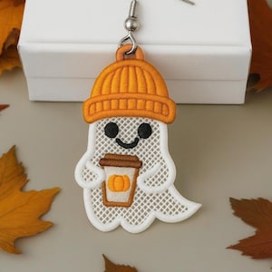 Puede incluir: Pendiente de fantasma bordado con gorro naranja y sosteniendo un café con leche de calabaza. El fantasma es blanco con un diseño de malla, ojos negros y boca sonriente. Accesorio con temática otoñal.
