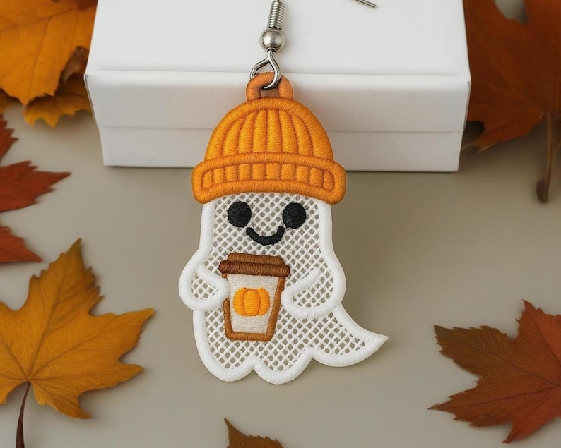 Puede incluir: Pendiente de fantasma bordado con gorro naranja y sosteniendo un caf&eacute; con leche de calabaza. El fantasma es blanco con un dise&ntilde;o de malla, ojos negros y boca sonriente. Accesorio con tem&aacute;tica oto&ntilde;al.