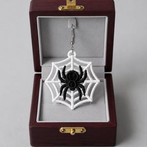 May include: A single earring featuring a black embroidered spider centered on a white embroidered spiderweb. The earring is displayed inside a dark wood jewelry box with a light gray interior lining and gold-toned metal hardware.