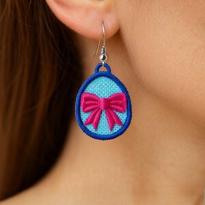 May include: A single earring with a blue oval design and a pink bow. The earring is embroidered with a blue border and a silver hook. The bow is a bright pink color.