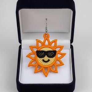 May include: An orange embroidered sun earring with black sunglasses is displayed inside a dark blue jewellery box. The sun has a smiling face and a yellow centre. The earring is hanging from a silver hook.