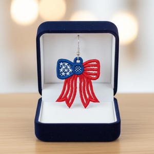 May include: A bow-shaped earring in a navy blue jewellery box. The earring features a blue centre with white stars and a red bow with red stripes, reminiscent of the American flag. The earring is displayed in a dark blue velvet box.