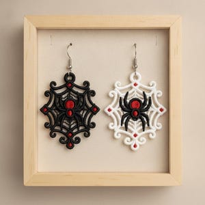 May include: A pair of dangling earrings featuring intricate embroidery of a spider centered on a web. One earring is black with a red spider body, and the other is white with a black and red spider. Both have silver-toned metal hooks.