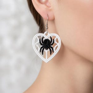 May include: A white embroidered heart-shaped earring featuring a black spider design in the center. The earring is suspended from a silver-toned metal hook and showcases intricate thread work forming a web pattern within the heart frame.