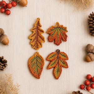 Op de afbeelding: Vier geborduurde bladornamenten in herfstkleuren zoals oranje, geel en groen. Elk blad is voorzien van gedetailleerd stikwerk en een klein lusje aan de bovenkant om op te hangen. De set bevat een eikenblad, een esdoornblad en twee andere bladvormen.